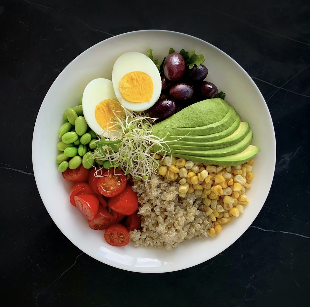 a bowl containing avocado, eggs, olives, quinoa, corn, tomatoes, and edamame