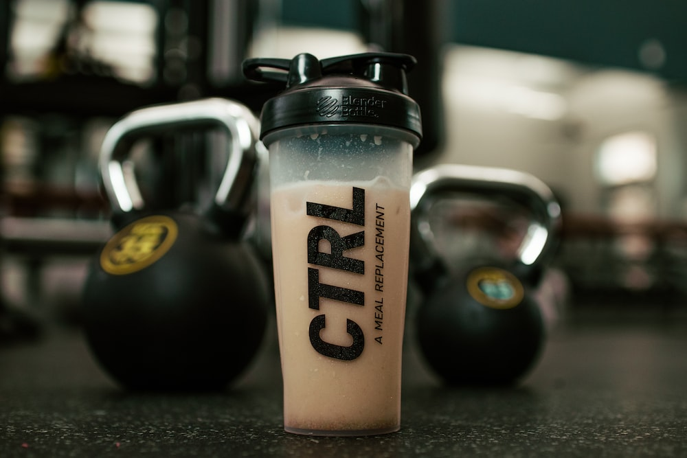 a meal replacement drink in a container with some kettlebells in the background
