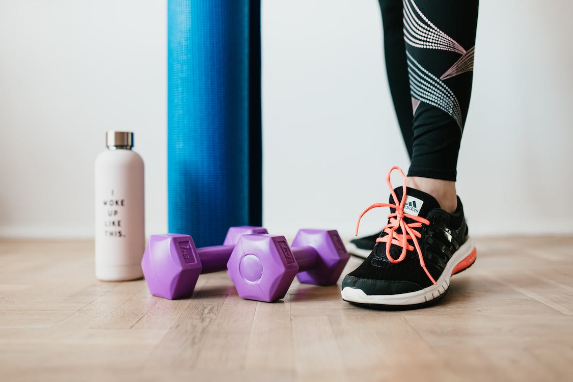 A person’s legs are seen next to two purple dumbbells, a blue yoga mat, and a white water bottle.